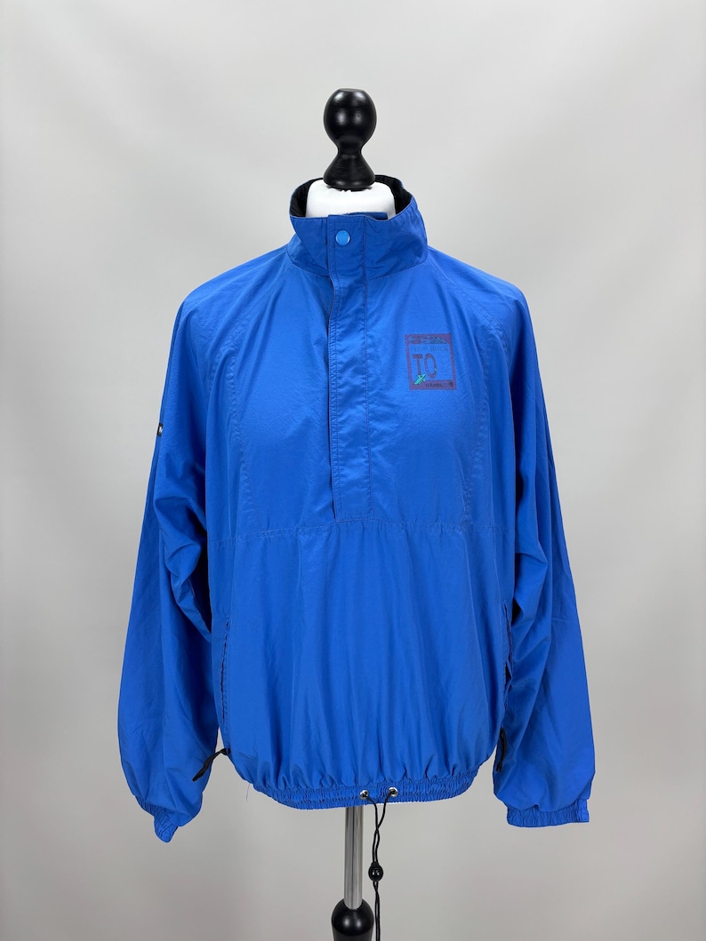 May include: Bright blue windbreaker jacket with a high collar and a quarter-zip closure. The jacket features a small logo on the chest and a drawstring at the hem. The sleeves are long and have elastic cuffs. The jacket is displayed on a mannequin.