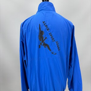 May include: Bright blue jacket with a high collar and a black graphic on the back that reads "ALPIN-SPORT GRABS" with a climber illustration. The jacket has a zipper closure and long sleeves. The jacket is displayed on a mannequin.
