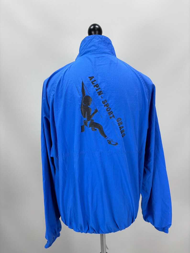 May include: Bright blue jacket with a high collar and a black graphic on the back that reads "ALPIN-SPORT GRABS" with a climber illustration. The jacket has a zipper closure and long sleeves. The jacket is displayed on a mannequin.