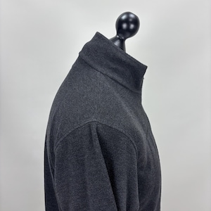 May include: A charcoal grey fleece pullover with a high collar, displayed on a black mannequin head. The fabric has a textured appearance. The background is plain white.