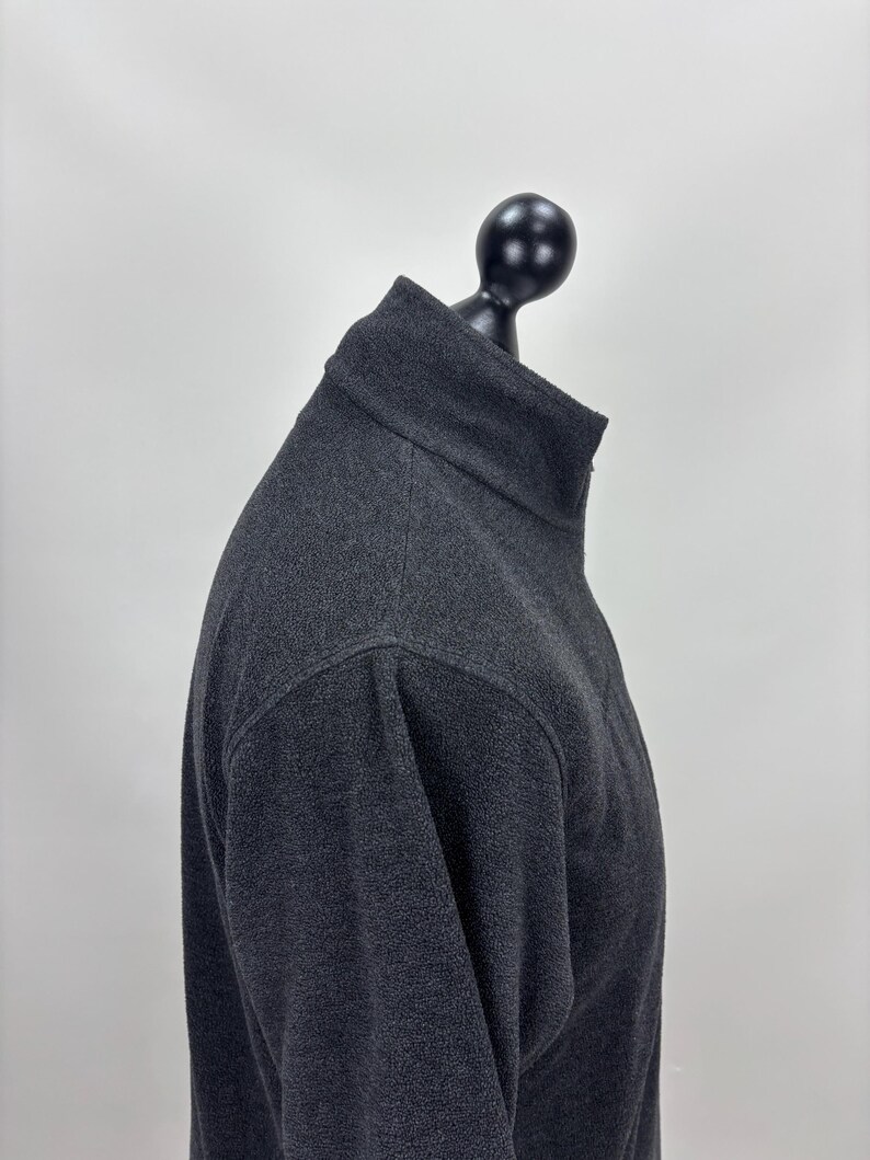 May include: A charcoal grey fleece pullover with a high collar, displayed on a black mannequin head. The fabric has a textured appearance. The background is plain white.