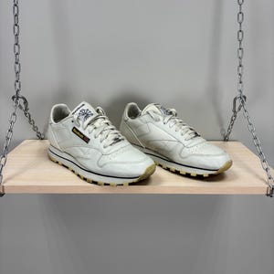 Op de afbeelding: Een paar off-white Reebok Classic Leather sneakers. De schoenen hebben een leren bovenwerk, een zwarte streep langs de tussenzool en een buitenzool in gumkleur. Het Reebok-logo is zichtbaar op de tong en zijkant. De sneakers staan op een houten plank.