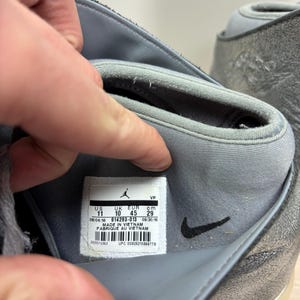 May include: Close-up of a gray and silver athletic shoe. The shoe's interior reveals a label with size information: US 11, UK 10, EUR 45, and 29 cm. The label also indicates the shoe was made in Vietnam.