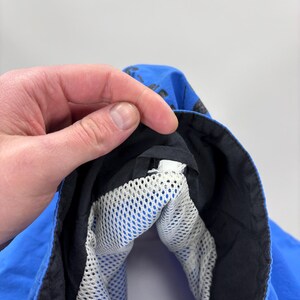 May include: Close-up of a blue jacket hood with a black trim and a white mesh lining. The hood is held open by a hand, revealing the interior construction and materials. The jacket appears to be made of a lightweight, water-resistant fabric.