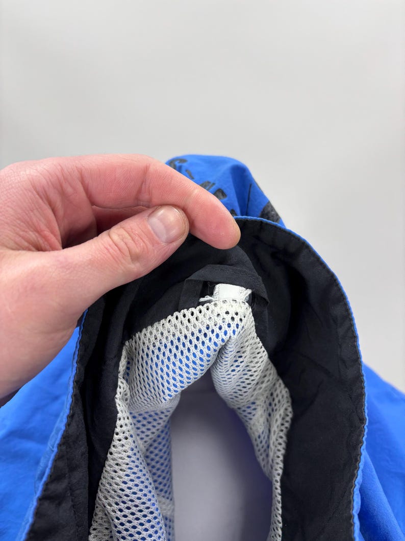 May include: Close-up of a blue jacket hood with a black trim and a white mesh lining. The hood is held open by a hand, revealing the interior construction and materials. The jacket appears to be made of a lightweight, water-resistant fabric.