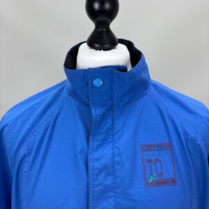 May include: Bright blue jacket with a black collar and a button closure at the neck. The jacket features a small rectangular logo on the right side with the text "IN A ROCK TO MAMMUT".