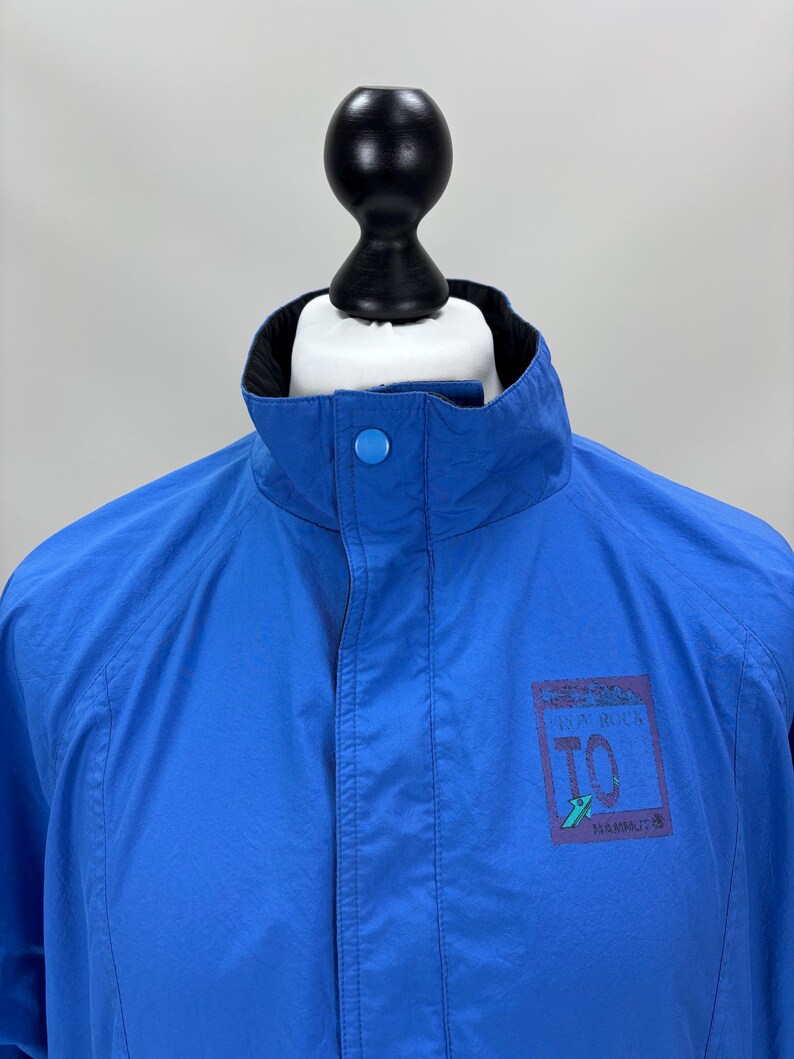 May include: Bright blue jacket with a black collar and a button closure at the neck. The jacket features a small rectangular logo on the right side with the text "IN A ROCK TO MAMMUT".