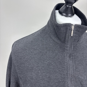 May include: A dark grey fleece jacket with a high collar and a full-length zip. The jacket features a small logo on the chest. The jacket is displayed on a mannequin.