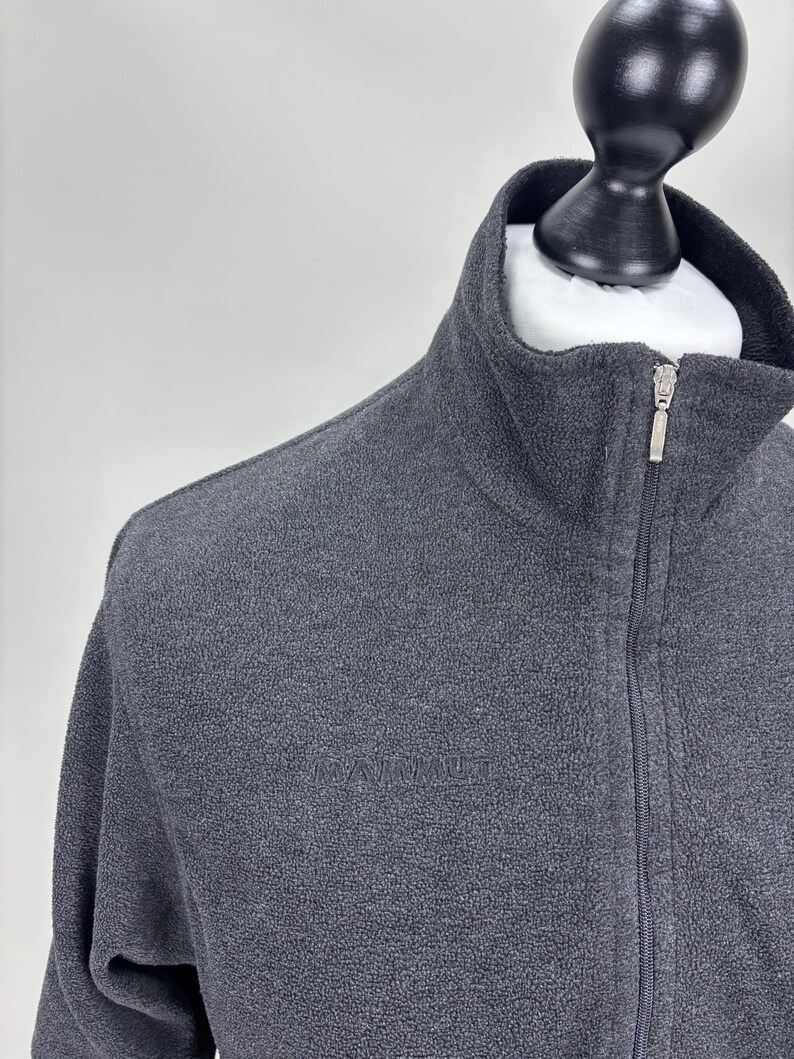 May include: A dark grey fleece jacket with a high collar and a full-length zip. The jacket features a small logo on the chest. The jacket is displayed on a mannequin.