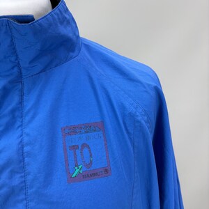 May include: Bright blue jacket with a high collar. The jacket features a small rectangular patch with the text "FROM ROCK TO" and the brand name "MAMMUT". The jacket is made of a lightweight material.