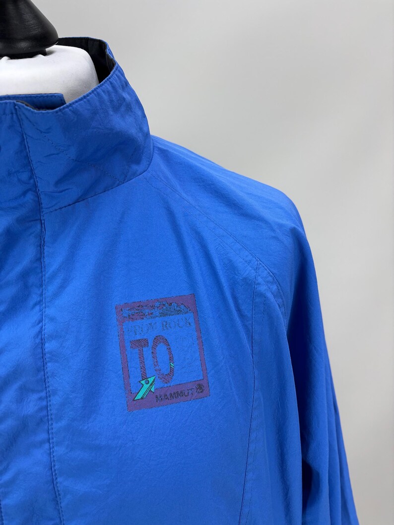 May include: Bright blue jacket with a high collar. The jacket features a small rectangular patch with the text "FROM ROCK TO" and the brand name "MAMMUT". The jacket is made of a lightweight material.