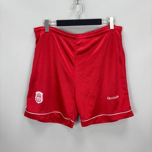May include: Red athletic shorts with white trim and a small embroidered emblem. The shorts feature the Reebok logo in white on the right leg. The shorts are hanging on a hanger.
