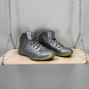May include: A pair of metallic silver high-top sneakers with gray laces and a yellowed sole. The shoes are displayed on a light wood shelf suspended by metal chains. The sneakers have a textured appearance and a classic athletic design.