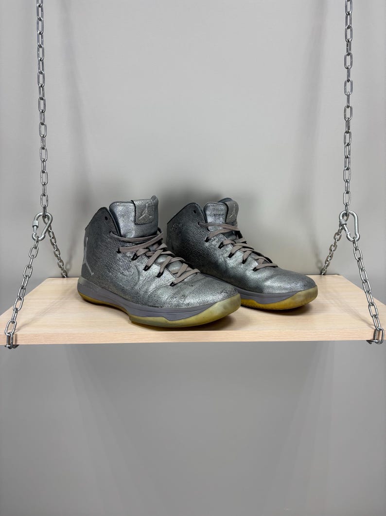 May include: A pair of metallic silver high-top sneakers with gray laces and a yellowed sole. The shoes are displayed on a light wood shelf suspended by metal chains. The sneakers have a textured appearance and a classic athletic design.