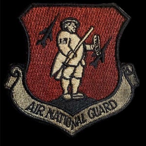 May include: A red and tan embroidered patch with a shield shape. The shield features a cartoon figure holding a weapon and a plane flying overhead. The text "AIR NATIONAL GUARD" is written in a banner below the shield.