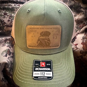 Puede incluir: Gorra de camionero verde oliva con parte trasera de malla beige. Cuenta con un parche de cuero con una ilustración de retriever y el texto: "MIGRATORY BIRD HUNTING STAMP". Incluye un parche Richardson que dice "ORIGINAL 112 TRUCKER ADJUSTABLE".