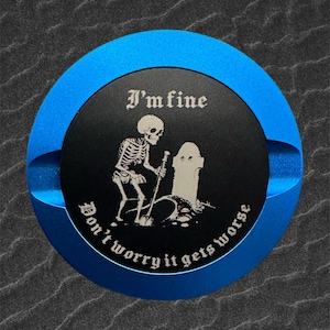 Puede incluir: Un objeto redondo azul y negro con los textos "I'm fine" y "Don't worry it gets worse". Presenta un esqueleto con una pala junto a una lápida.