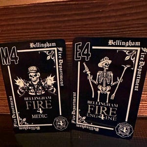 May include: Two black playing cards with white illustrations of skeletons in firefighter uniforms. One card says "M4 Bellingham Fire Medic" and the other says "E4 Bellingham Fire Engine 4". Both cards display the Bellingham Fire Department logo.