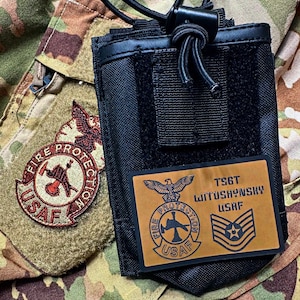 May include: Black tactical radio pouch with a hook and loop closure and drawstring. A brown patch with the text "FIRE PROTECTION USAF" and a second patch with the text "TSGT WITOSHYNSKY USAF" are attached to the camouflage fabric.