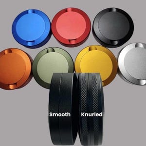 Op de afbeelding: Een verzameling kleurrijke, ronde metalen objecten in blauw, rood, zwart, oranje, olijfgroen, goud en zilver. Twee zwarte cilindrische objecten eronder, één glad en één gekarteld, met de woorden "Smooth" en "Knurled" erop.