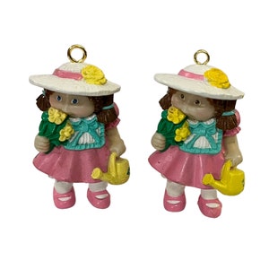 Puede incluir: Dos adornos de Cabbage Patch Kid, cada uno con un sombrero de sol blanco con una cinta rosa y sosteniendo un ramo de flores amarillas y una regadera amarilla.