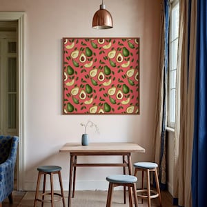 Könnte beinhalten: Ein gerahmtes Kunstwerk mit einem sich wiederholenden Muster aus Avocados und Blättern auf korallenfarbenem Hintergrund. Der Raum umfasst einen Holztisch, drei Hocker mit blauen Sitzen und eine blaue Vase mit einer kleinen Pflanze.