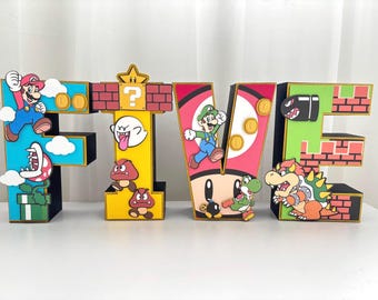 May include: Five wooden letters decorated with characters from the Super Mario video game franchise. The letters are painted in red, yellow, green, and blue with gold accents. The letters are decorated with characters such as Mario, Luigi, Bowser, and Goombas.
