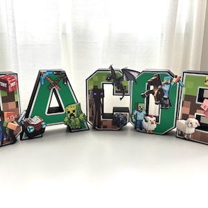 Può includere: Lettere con nome a tema Minecraft che compongono "JACOB". Ogni lettera è di un colore diverso e presenta personaggi ed elementi di Minecraft, tra cui un Creeper, un Enderman e un maiale. Le lettere sono su una superficie bianca.