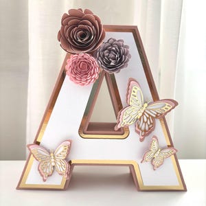 Op de afbeelding: Een decoratieve letter "A" in een gelaagd ontwerp, met een witte binnenkant, een roségouden rand en gouden accenten. Versierd met papieren bloemen in roze en mauve, en delicate vlinderuitsparingen.