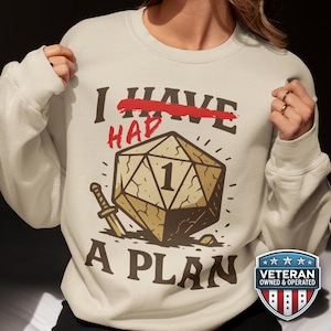 May include: Cream-colored sweatshirt with the text "I HAVE HAD A PLAN" and a large die graphic. The word "HAVE" is crossed out in red. A small sword graphic is next to the die. A Veteran Owned & Operated badge is on the bottom right.