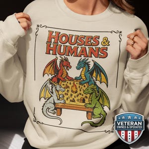 Puede incluir: Sudadera color crema con un gráfico de cuatro dragones alrededor de una mesa y el texto "HOUSES & HUMANS". Los dragones son rojos, azules, verdes y grises. Un escudo "Veteran Owned & Operated" está en la parte inferior.