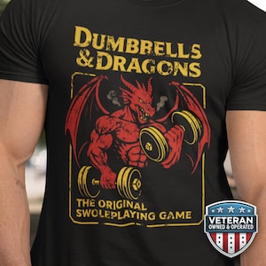 May include: Black t-shirt with a yellow and red graphic. The graphic features a red dragon lifting yellow dumbbells, with the text "Dumbbells & Dragons" and "The Original Swoleplaying Game". A "Veteran Owned & Operated" badge is on the bottom right.