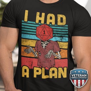 May include: Black t-shirt with a distressed graphic. The design features the text "I HAD A PLAN" in yellow, with a skeleton figure wearing a red D20 die as a head. The design has a retro, vintage style.
