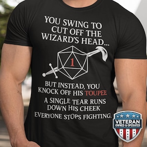 Puede incluir: Camiseta negra con texto blanco y un gráfico de dado, espada y hacha. El texto dice: "YOU SWING TO CUT OFF THE WIZARD'S HEAD... BUT INSTEAD, YOU KNOCK OFF HIS TOUPEE." Una insignia de "Veteran Owned & Operated" está en la parte inferior derecha.