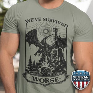 May include: Sage green t-shirt with a black graphic of a dragon, castle, and the text "WE'VE SURVIVED WORSE." A Veteran Owned & Operated badge is on the lower right.