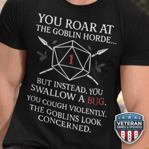 May include: Black t-shirt with white text and graphics. The text reads "YOU ROAR AT THE GOBLIN HORDE... BUT INSTEAD, YOU SWALLOW A BUG. YOU COUGH VIOLENTLY. THE GOBLINS LOOK CONCERNED." A D20 die is in the center.