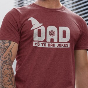 以下が含まれることがあります： 霜降りマルーンカラーの半袖Tシャツで、白いグラフィックプリントが施されています。デザインは「DAD」という文字の上に魔法使いの帽子、Aの文字の中に20面ダイス、そしてその下に白い枠で囲まれた「+5 TO DAD JOKES」というテキストが配置されています。