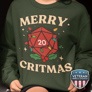 May include: A dark green sweatshirt with the words "MERRY CRITMAS" in a cream color. The design features a red D20 die with the number 20, decorated with Christmas lights and holly.