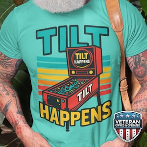 Peut inclure: T-shirt turquoise avec un motif de flipper coloré. Le design présente les mots "TILT HAPPENS" en lettres grasses soulignées. Le flipper est rouge avec des accents jaunes et bleus. Le t-shirt a un style rétro.