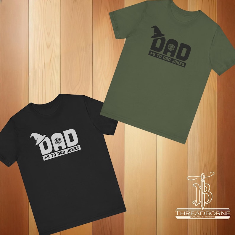 以下が含まれることがあります： 黒とオリーブグリーンの半袖Tシャツ2枚。それぞれに「DAD +5 TO DAD JOKES」というグラフィックプリントがあり、魔法使いの帽子のアイコンが「A」の文字を置き換え、20面ダイスのアイコンが「A」の文字の中に配置されています。