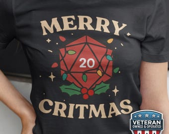 以下が含まれることがあります： お祝いのデザインが施された黒いTシャツ。「MERRY CRITMAS」の文字が、赤いD20サイコロの上下に配置されています。サイコロには20の数字が入り、クリスマスライトとヒイラギで飾られています。「Veteran Owned & Operated」のバッジが右下にあります。