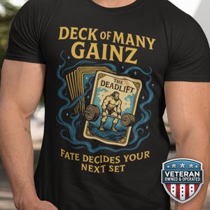 May include: Black t-shirt with a graphic featuring a weightlifter on a card labeled "The Deadlift." Text reads "Deck of Many Gainz" and "Fate Decides Your Next Set." A "Veteran Owned & Operated" badge is on the bottom right.