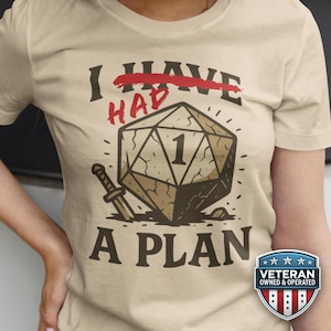 Könnte beinhalten: Beiges T-Shirt mit einer Grafik, die einen großen Würfel mit der Zahl 1, ein Schwert und den Text "I HAD A PLAN" zeigt. Das Wort "HAD" ist rot durchgestrichen. Ein "Veteran Owned & Operated"-Abzeichen befindet sich unten rechts.