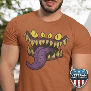 May include: Rust-colored t-shirt featuring a cartoon monster face with yellow eyes, sharp teeth, and a purple tongue. The design is centered on the shirt. A Veteran Owned & Operated badge is on the lower right.