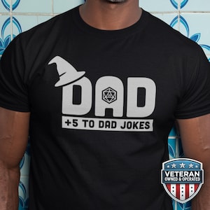 以下が含まれることがあります： 黒の半袖Tシャツで、白いグラフィックがプリントされています。DADという文字の最初の文字の上に魔法使いの帽子があり、Aの文字の中に20面ダイスが描かれています。その下に「+5 TO DAD JOKES」というテキストがあります。退役軍人所有のロゴが表示されています。