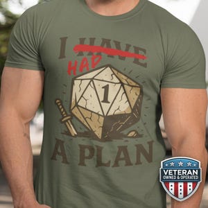 May include: Olive green t-shirt with a graphic featuring a large die with the number 1, a sword, and the text "I HAD A PLAN". The text "HAVE" is crossed out in red. A "Veteran Owned & Operated" badge is on the bottom right.