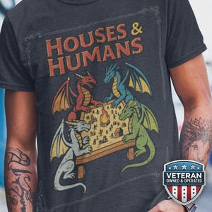 Może przedstawiać: Ciemnoszary t-shirt z nadrukiem graficznym z napisem "HOUSES & HUMANS" pomarańczowymi literami. Wzór przedstawia cztery smoki w kolorach czerwonym, niebieskim, zielonym i szarym, zgromadzone wokół drewnianego stołu podczas gry w planszówkę.