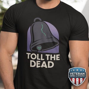Pode incluir: Camiseta preta com um gráfico de um sino com uma caveira e o texto "TOLL THE DEAD". O sino é cinza escuro com um design de raio verde. Um distintivo "Veteran Owned & Operated" está no canto inferior direito.