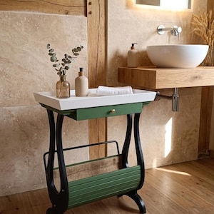 Może przedstawiać: Dekoracyjny stolik boczny do łazienki z białym blatem, zieloną szufladą i czarnymi nogami. Stół posiada stojak na czasopisma i mieści ręcznik, dozownik mydła oraz wazon z zielenią. Stół wykonany jest z drewna i metalu.