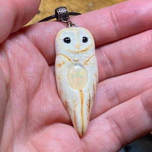 May include: A pendant shaped like a barn owl with a cream-colored body, brown feather details, and small black eyes. An oval-shaped iridescent opal cabochon is embedded in the center of the owl's chest. The pendant hangs from a black cord.
